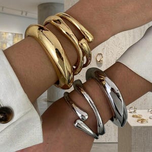 Könnte beinhalten: Ein Set aus drei goldfarbenen Metallarmreifen mit glatter, polierter Oberfläche an einem Arm und ein Set aus drei silberfarbenen Metallarmreifen mit passender glatter, polierter Oberfläche am anderen Arm, beide mit offenem Design.