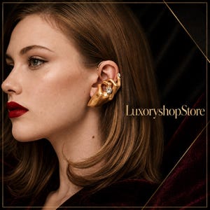 Peut inclure: Une boucle d'oreille en forme de main dorée, ornée d'une petite pierre transparente sur le doigt et d'une autre à l'extrémité. Le bijou s'enroule autour du cartilage de l'oreille. Le texte LuxoryshopStore est visible sur l'image.