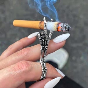 Op de afbeelding: Een zilverkleurige metalen ring in de vorm van een skeletachtige hand met uitgestrekte vingers, ontworpen om een brandende sigaret vast te houden. De ring heeft gedetailleerde botstructuren en gewrichten die het sigarettenfilter stevig vasthouden.