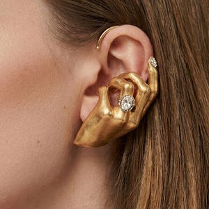 Puede incluir: Un pendiente tipo ear cuff dorado con forma de mano que rodea la oreja. La mano presenta una pequeña piedra ovalada transparente engastada en un anillo en el dedo corazón y un detalle de piedra transparente en la punta del dedo índice.