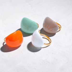 Puede incluir: Cuatro anillos coloridos con piedras redondeadas en forma de lágrima. Los anillos presentan bandas doradas y están en tonos naranja, verde menta, blanco y marrón claro. Se exhiben sobre una superficie gris claro.