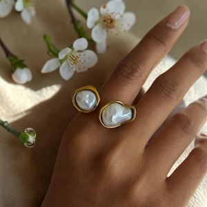 May include: A gold-toned open ring featuring two irregular, organic-shaped white pearls. The ring band is smooth and metallic, holding one smaller pearl and one larger pearl in a bypass style, creating a distinct contrast between the metal and pearls.