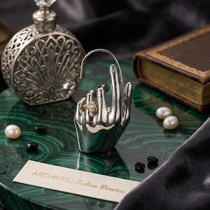 May include: A silver-toned hand-shaped jewellery display stand featuring a gold-toned ring with a clear stone on the ring finger. A small rectangular card with the text ARCHIVES | Fashion Direction is placed in front of the display stand.