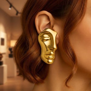 May include: A gold-toned earring featuring a sculpted, abstract human face design with smooth, reflective surfaces. The piece covers the earlobe and extends upward, showcasing detailed contours of closed eyes, a nose, and lips in a metallic finish.