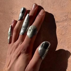 Puede incluir: Un conjunto de cuatro piezas de armadura de dedo de metal plateado que se usan en los dedos. Cada pieza presenta un acabado metálico texturizado que cubre la punta del dedo y el área de la uña, extendiéndose una de ellas como un anillo.
