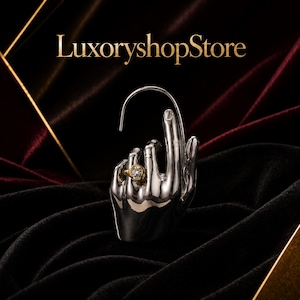 Pode incluir: Uma escultura de mão metálica prateada com um anel dourado com uma pedra clara no dedo anelar. O texto LuxoryshopStore é exibido acima da escultura. A mão está posicionada com os dedos curvados e um fio fino arqueado por cima.