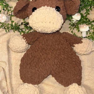 May include: A handmade brown and cream-colored crochet cow plushie. The plush toy has a brown body, ears, and curly hair, with a cream-colored muzzle, paws, and feet. The cow has black safety eyes.