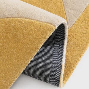 May include: A rolled-up area rug with a geometric design. The rug features a mustard yellow, cream, and light gray color scheme. The underside of the rug is a dark gray with a white herringbone pattern.
