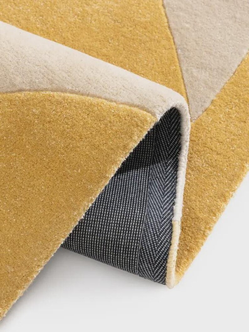 May include: A rolled-up area rug with a geometric design. The rug features a mustard yellow, cream, and light gray color scheme. The underside of the rug is a dark gray with a white herringbone pattern.