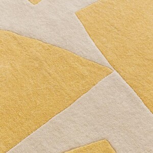 May include: Close-up of a geometric rug with a repeating pattern. The rug features a combination of mustard yellow and off-white colors. The design consists of angular shapes and lines, creating a modern aesthetic.