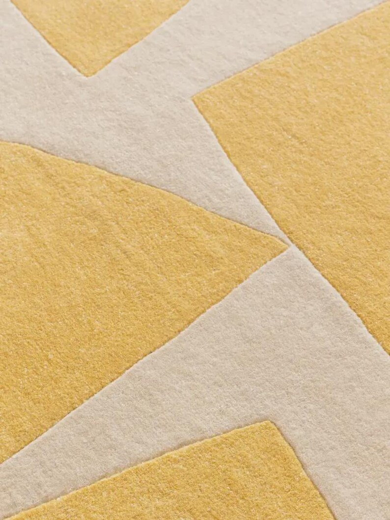 May include: Close-up of a geometric rug with a repeating pattern. The rug features a combination of mustard yellow and off-white colors. The design consists of angular shapes and lines, creating a modern aesthetic.