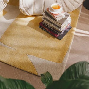 May include: A yellow and beige patterned rug with a stack of books and a teacup on top. The rug has a geometric design with a soft, textured appearance. A white, cushioned chair is in the background.