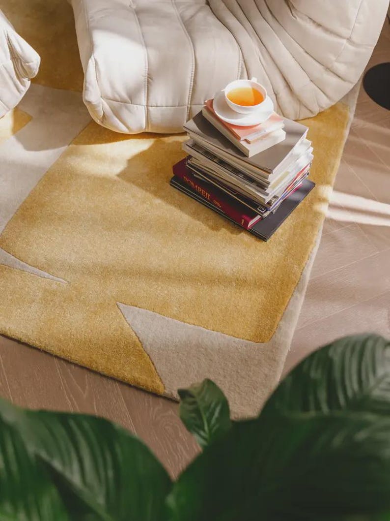May include: A yellow and beige patterned rug with a stack of books and a teacup on top. The rug has a geometric design with a soft, textured appearance. A white, cushioned chair is in the background.