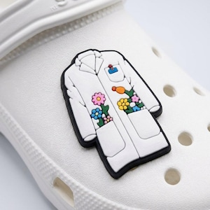 May include: A white shoe with a decorative charm shaped like a lab coat. The lab coat charm has black trim and is decorated with colorful flowers and a small badge. The shoe is white.