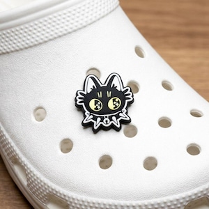 May include: A black cat-shaped shoe charm with yellow eyes and a spiky collar is attached to a white shoe. The charm has a cartoon-like design and is made of a rubbery material. The shoe has several round holes.