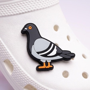 Puede incluir: Un adorno para zapatos con forma de paloma, con un diseño negro, gris y blanco, y patas y ojo naranjas. El adorno está adherido a un zapato blanco, con agujeros visibles.