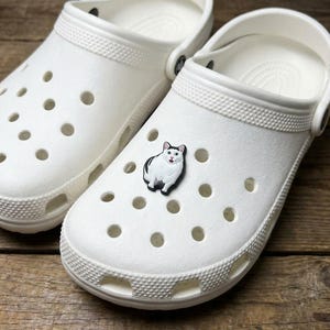 May include: White clogs with a cat-shaped charm. The clogs are made of a smooth, solid white material with multiple ventilation holes and a textured band around the top. The cat charm is black and white.