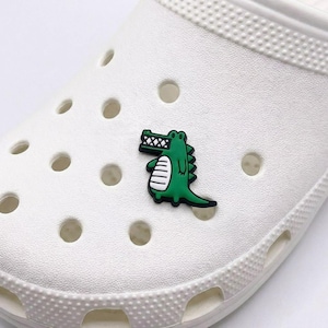 May include: A green and black alligator shoe charm is attached to a white shoe. The alligator has a detailed design with white stripes on its belly and a textured back. The shoe has multiple holes.