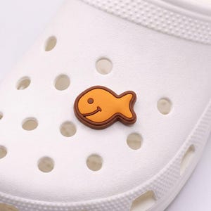 Könnte beinhalten: Ein weißer Clog-Schuh mit einem dekorativen orangefarbenen und braunen Fisch-Charm. Der Fisch-Charm lächelt und hat eine braune Umrandung. Der Schuh hat mehrere kreisförmige Belüftungslöcher.