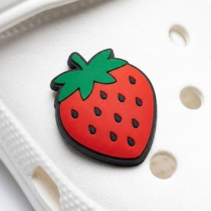 May include: A red and black strawberry shoe charm with a green stem and leaves. The charm has black seeds and is set against a white background with two round holes.