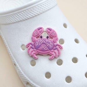 May include: A pink and purple crab shoe charm is attached to a white shoe. The crab charm has a detailed design with a lighter pink outline and a darker purple center. The shoe has multiple round holes.