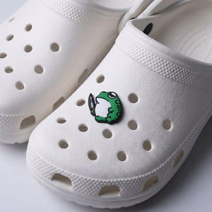 May include: White clogs with a green frog charm holding a knife. The frog charm is green, white, and black. The clogs have multiple ventilation holes and a textured surface.