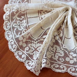 May include: Cream-colored, vintage lace tablecloth with intricate openwork and a scalloped edge. The detailed design features geometric patterns and floral motifs, creating an elegant and delicate aesthetic. The fabric is folded, showcasing the texture and craftsmanship.