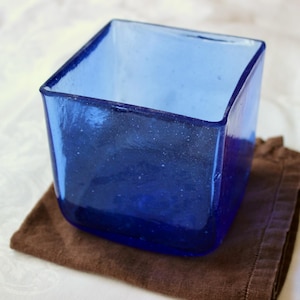 May include: A square, cobalt blue glass container with rounded corners. The container sits on a brown, textured cloth. The glass has a slightly uneven surface, giving it a handmade appearance. The container is empty.