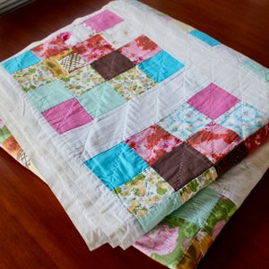 May include: A handmade quilt with a patchwork design. The quilt features squares of various colors, including pink, blue, brown, and floral patterns, set against a white background. The quilt is folded, showing the detailed stitching and layered construction.