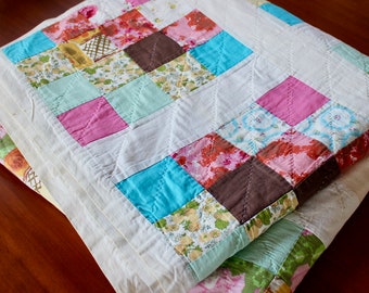 May include: A handmade quilt with a patchwork design. The quilt features squares of various colors, including pink, blue, brown, and floral patterns, set against a white background. The quilt is folded, showing the detailed stitching and layered construction.