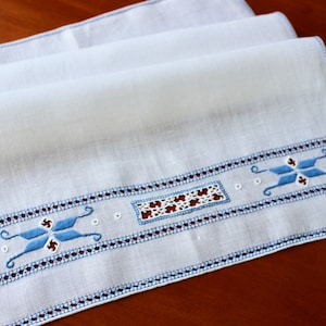 May include: White linen table runner with blue embroidered details. The embroidery features a geometric pattern with a central star shape.