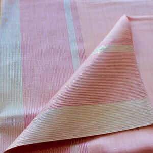 May include: Close-up of a pink fabric with vertical stripes in light pink and silver. The fabric is folded over, revealing the texture and sheen of the material. The overall aesthetic is soft and elegant.