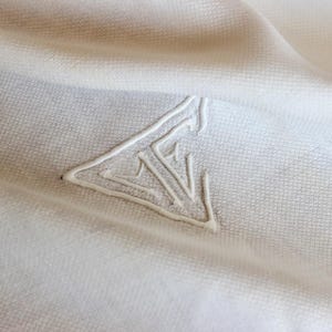 May include: A white fabric with a subtle, repeating grid-like texture featuring a raised, embroidered triangular logo in the center. The design consists of white thread forming a geometric shape with internal lines, creating a monochromatic look.