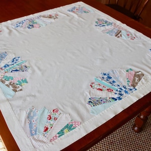May include: A white tablecloth with colorful, fan-shaped patchwork designs in each corner. The patchwork includes floral, paisley, and striped patterns in shades of blue, pink, and green. The tablecloth is on a dark wooden table.