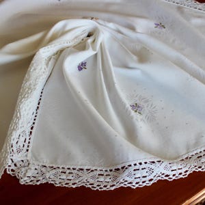 May include: Ivory-colored tablecloth with delicate floral embroidery and a crocheted lace border. The fabric is draped, showcasing the intricate details of the design. Small purple flower clusters are scattered across the cloth.