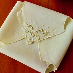May include: A folded, pale yellow linen handkerchief with a delicate floral embroidery in the corner. The embroidery features a cut-out design with small, raised dots and a scalloped edge. The handkerchief is neatly folded, showcasing the detailed craftsmanship.