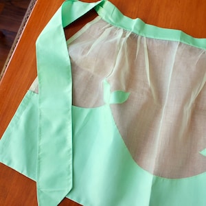 May include: A mint green half apron with a sheer, semi-transparent overlay. The apron features a solid mint green waistband and ties, with decorative leaf-shaped cutouts on the lower portion. The apron is made of a lightweight fabric.