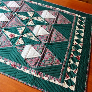 May include: A green quilted table runner with a paisley pattern and white triangles. The runner has a border of green and white triangles.