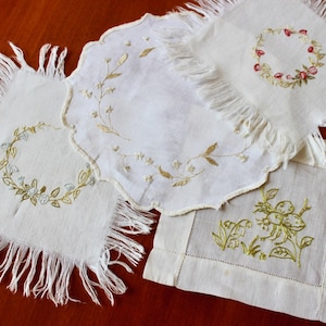 May include: A collection of four white linen coasters with embroidered floral designs. The coasters feature fringed edges and various botanical patterns in gold, green, and red thread, including wreaths and sprigs.