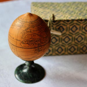 May include: A small, antique-style globe with a light brown surface, featuring detailed black line drawings of continents and oceans. The globe sits on a dark green, circular base. A patterned box is in the background.