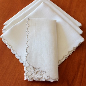 May include: A stack of white linen napkins with scalloped edges and a floral embroidery design. The napkins are neatly folded, showcasing the delicate details of the fabric and embroidery. The napkins are on a wooden surface.