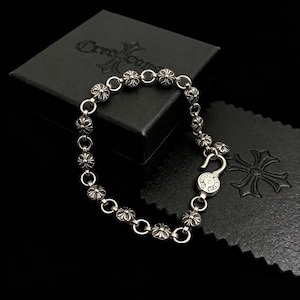 May include: A silver bracelet with a floral design, featuring a hook clasp. The bracelet is composed of small, round, flower-shaped beads connected by circular links. The bracelet rests on a black surface next to a black box with a logo.