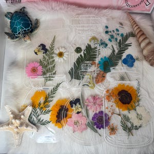 May include: Clear acrylic jar-shaped art pieces filled with dried flowers, including sunflowers, daisies, and pink blooms. A starfish, a sea turtle figurine, and a seashell are placed around the art pieces on a white fluffy surface.