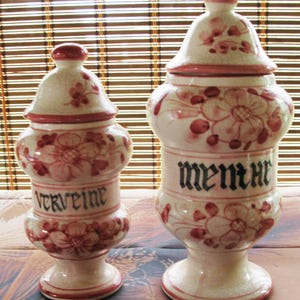 Könnte beinhalten: Zwei Keramik-Gläser mit Deckeln, verziert mit rosa Blumen und schwarzen Schriftzügen. Das Glas links trägt die Aufschrift "Verveine" und das Glas rechts die Aufschrift "Menthe".