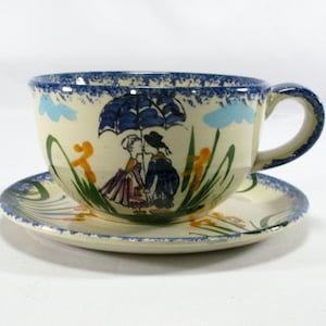 Puede incluir: Juego de taza y platillo de té de cerámica color crema. La taza presenta un borde azul y una escena pintada de dos figuras bajo un paraguas. El platillo tiene un diseño a juego. La taza mide unos 10 cm de altura.