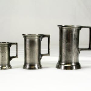 May include: Three silver measuring cups with handles. The smallest cup is labeled "1/2 LITRE", the middle cup is labeled "1 LITRE", and the largest cup is unlabeled.