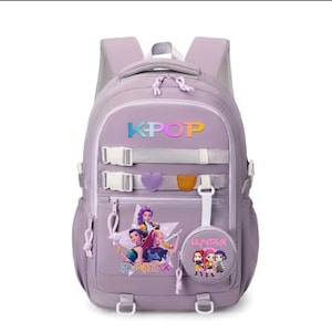 Op de afbeelding: Een lichtpaarse rugzak met de tekst KPOP in een verloopkleur, twee horizontale gespriemen, een paarse hartvormige patch en een rond tasje aan de voorzijde. De tas toont een afbeelding van drie geanimeerde personages en de tekst HUNTRAX.