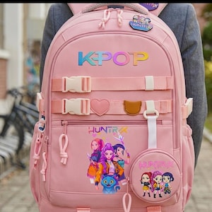 Puede incluir: Una mochila rosa con el texto KPOP en degradado, dos correas horizontales con hebillas de plástico, un parche en forma de corazón, un parche marrón y un estuche redondo. El frente muestra un gráfico de tres personajes y el texto HUNTRIX.