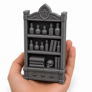 May include: A gray miniature bookshelf featuring three shelves filled with small potion bottles, stacked books, and a skull. The top of the unit is arched with a carved pentagram symbol. The base includes two small drawers with handles.