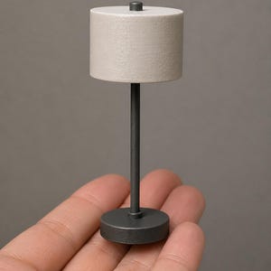 May include: Miniature table lamp with a cylindrical off-white lampshade and a dark gray metal base and stem. The lamp is a small scale model, likely for a dollhouse or miniature scene.